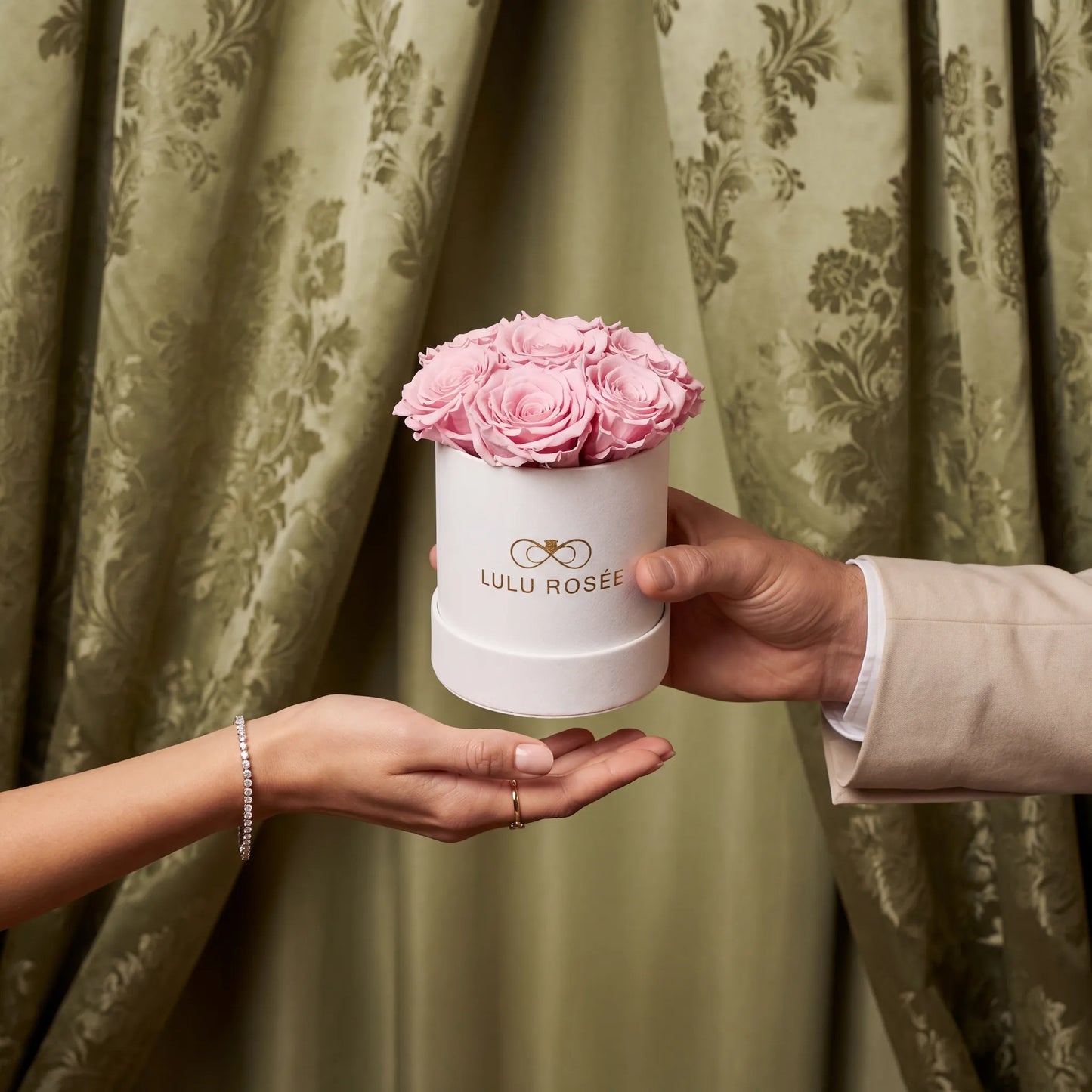 Person holding a small white box with pink roses against a green curtain background