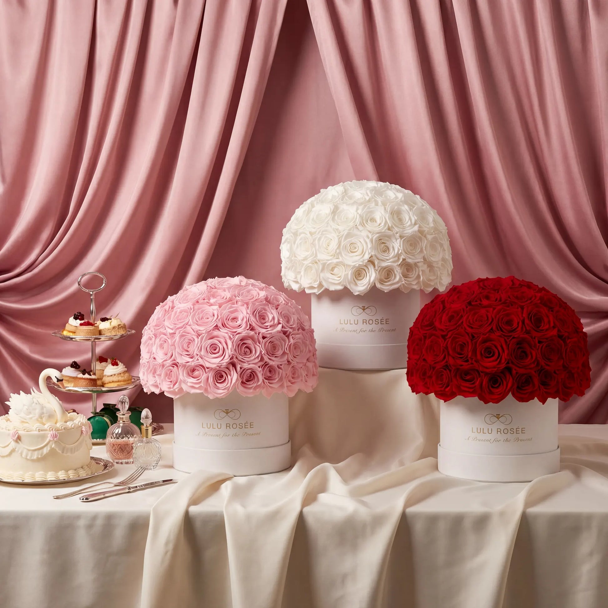 Three floral boxes in pink, white, and red with a cake and pastries on a table against a pink curtain.