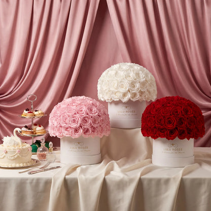 Three floral boxes in pink, white, and red with a cake and pastries on a table against a pink curtain.