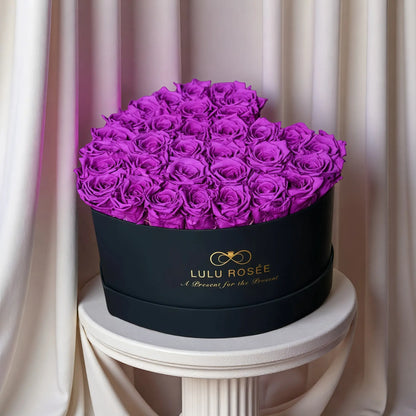 LULU ROSÉE large heart box purple roses black classic box on a white pedestal against a beige curtain background