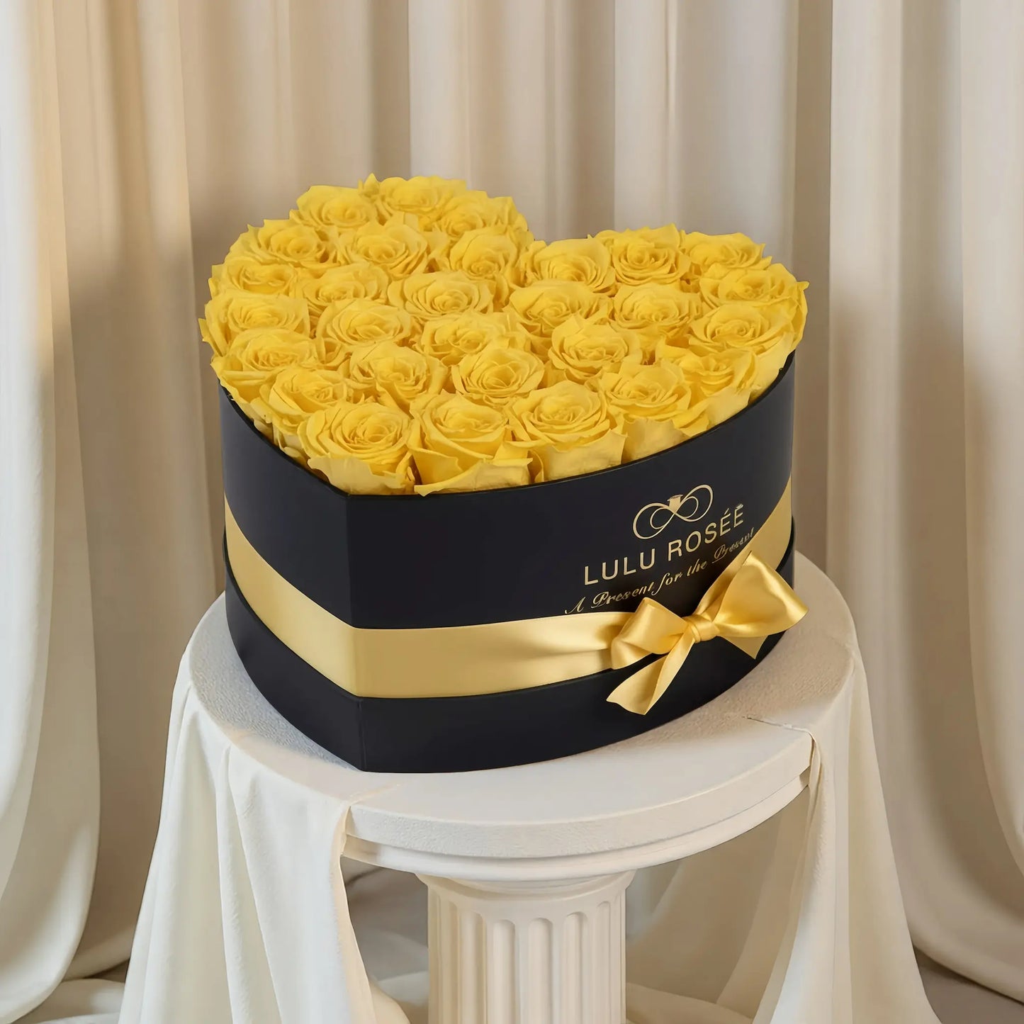 LULU ROSÉE large heart box yellow roses black classic box on a white pedestal against a beige curtain background