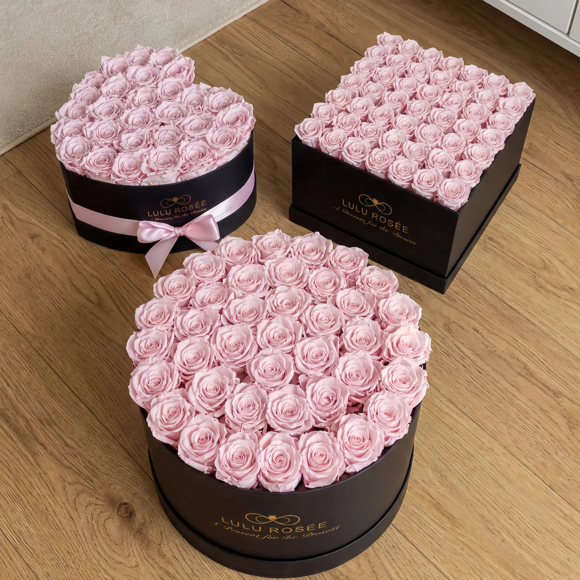 Three black boxes filled with pink roses on a wooden floor, branded 'Lulu Rosee'.